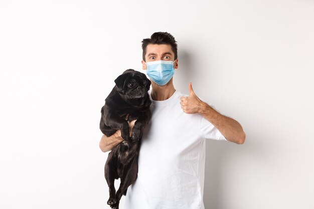 Covid-19, animals and quarantine concept. Young man in medical mask holding cute black pug dog, showing thumb up, like and approve, standing over white background