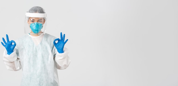 Free photo covid-19, coronavirus disease, healthcare workers concept. confident and serious asian female doctor, lab technician in personal protective equipment showing okay gesture in approval