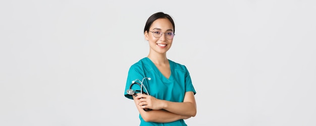 Covid-19, coronavirus disease, healthcare workers concept. Professional good-looking asian doctor, medical worker in glasses and scrubs, cross arms and smiling, white background.