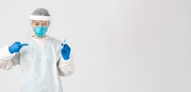 Covid-19, coronavirus disease, healthcare workers concept. Serious asian female doctor, physician in PPE personal protective equipment, pointing finger at syringe with vaccine, white background