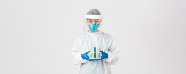 Covid-19, coronavirus disease, healthcare workers concept. Surprised asian female doctor, tech lab in personal protective equipment holding test-tubes with vaccine or analyses, white background.