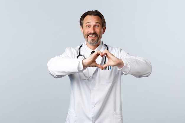 Free photo covid-19, coronavirus outbreak, healthcare workers and pandemic concept. happy caring male doctor in white coat, showing heart sign and smiling, express sympathy and admiration