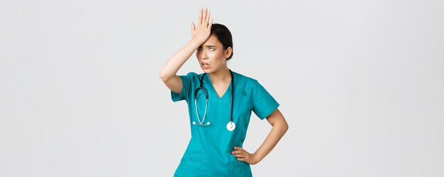 Covid-19, healthcare workers, pandemic concept. Annoyed and pissed-off asian female doctor in scrubs slap forehead, facepalm and eyeroll from someone stupid, tired standing white background.