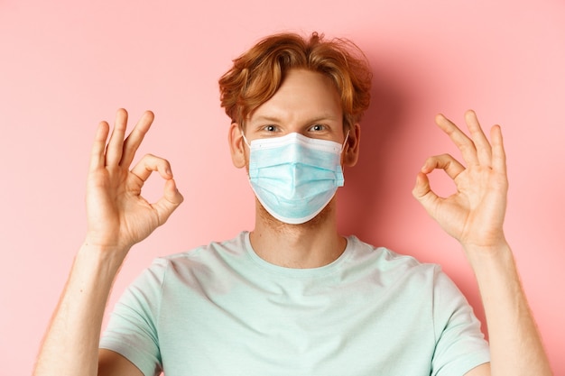 Free photo covid-19 and pandemic concept. handsome guy with messy ginger hair, wearing medical mask on face and showing okay signs, standing over pink background.