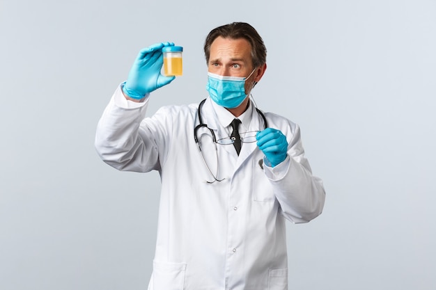 Covid-19, preventing virus, healthcare workers and vaccination concept. Serious-looking doctor in medical mask and gloves examine urine sample, look thoughtful and concentrated