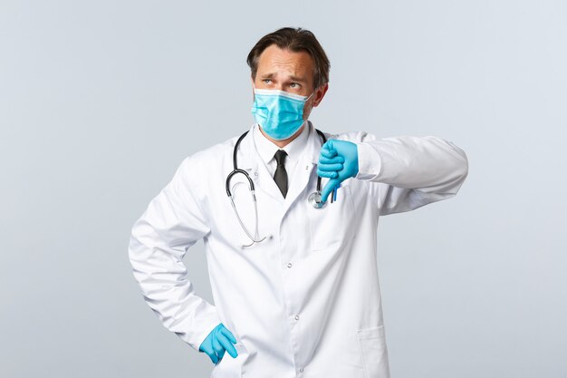 Covid-19, preventing virus, healthcare workers and vaccination concept. Skeptical and disappointed doctor in medical mask, gloves show thumb-down displeased, look top left sad