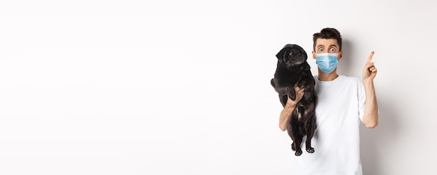 Covid animals and quarantine concept young man in medical mask holding cute black pug dog looking ri