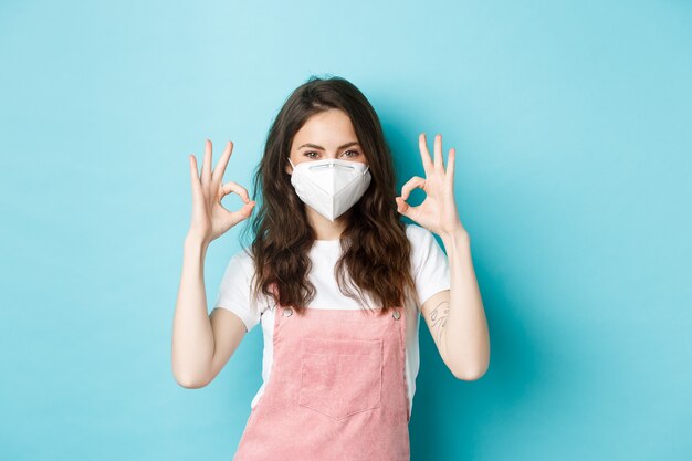 Covid, health and pandemic concept. Satisfied beautiful girl in respirator, medical mask showing okay sign in approval, uisng preventing measures from coronavirus, blue background.