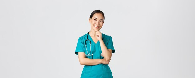 Covid healthcare workers pandemic concept smiling pleased attractive asian female doctor in scrubs l...