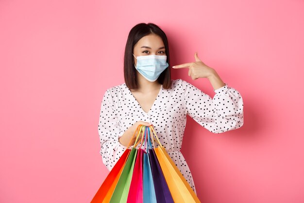 Covid pandemic and lifestyle concept cute asian woman holding shopping bags smiling and pointing at ...