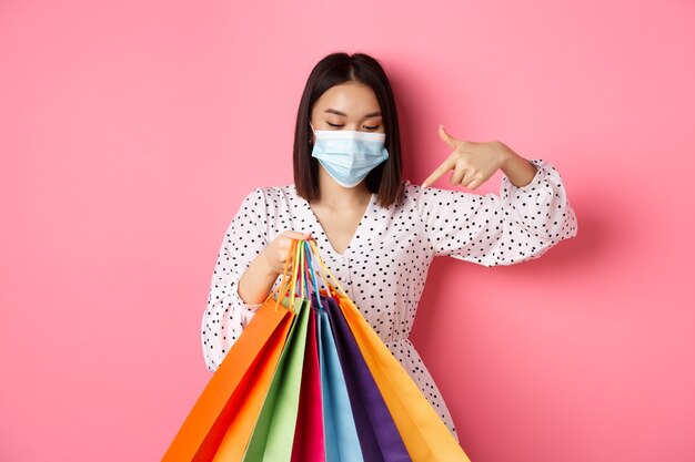 Covid pandemic and lifestyle concept pretty korean woman in medical mask pointing at shopping bags b...