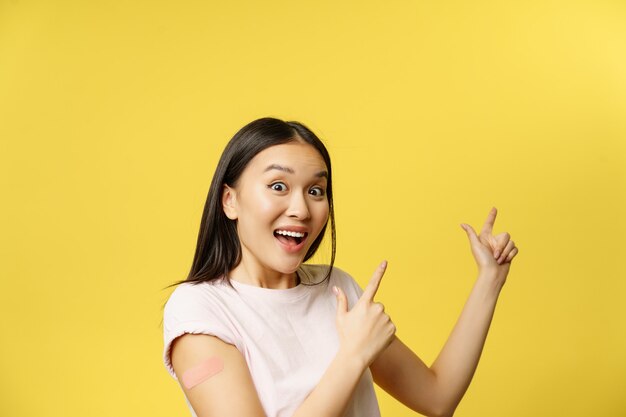 Covid vaccination concept happy vaccinated korean girl with patch on arm pointing at promo banner sh...