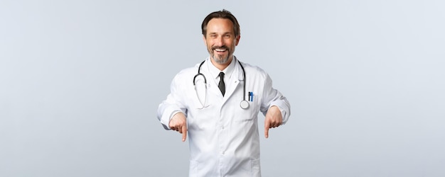 Free photo covid19 coronavirus outbreak healthcare workers and pandemic concept cheerful smiling male doctor in white coat inviting take test in clinic pointing fingers down advertising