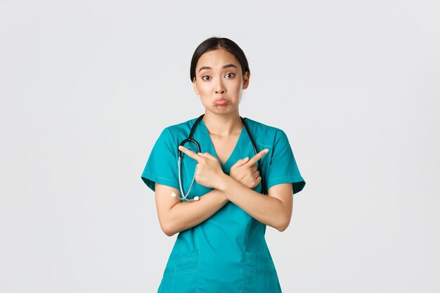 Covid19 healthcare workers pandemic concept Indecisive and clueless asian female nurse intern dont know what choose pointing fingers sideways and shrugging unaware white background