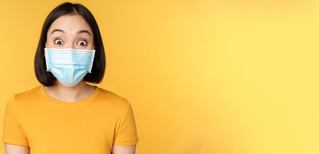 Covid19 and medical concept Close up portrait of asian woman in face mask looking surprised and amazed at news standing over yellow background