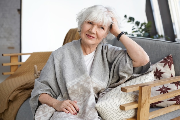 Coziness, interior design, leisure, lifestyle and elderly people concept. Attractive elegant mature female pensioner with wrinkles and gray hair relaxing on sofa in her country house, smiling