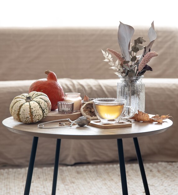 Cozy home still life with a cup of tea, pumpkins, candles and autumn decor details on a table on a blurred background of the room.