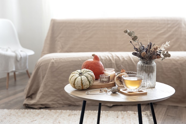 Cozy home still life with a cup of tea, pumpkins, candles and autumn decor details on a table on a blurred background of the room.