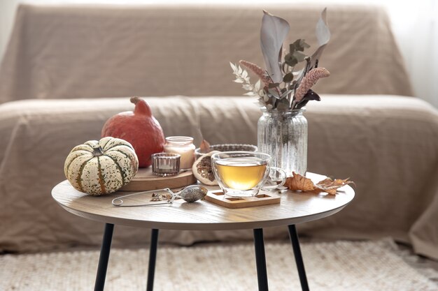 Cozy home still life with a cup of tea, pumpkins, candles and autumn decor details on a table on a blurred background of the room.