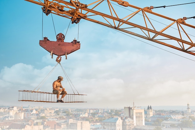 Free photo crane holding iron construction, where sitting builder with bare torso, eating and drinking milk. extreme building on high. cityscape on background.