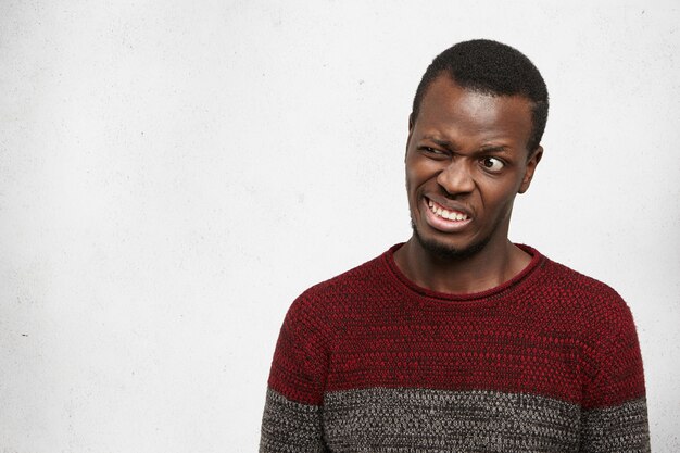 crazy funny young African American man wearing casual sweater posing indoors grimacing, making mouths, clenching teeth and winking. Human facial expressions, emotions and feelings