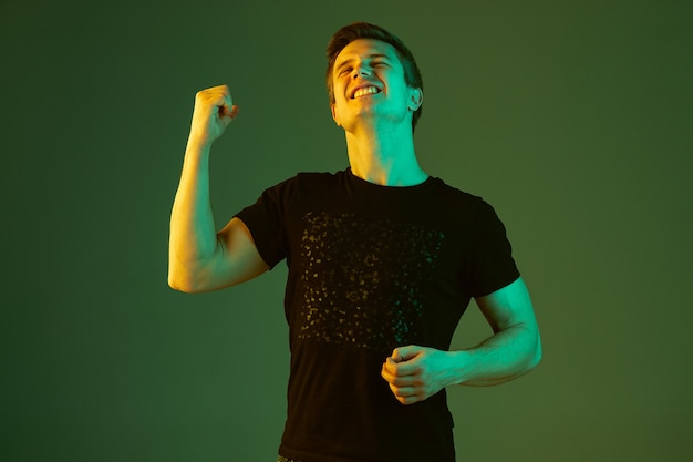 Crazy happe, celebrates win. Caucasian man's portrait isolated on green studio background in neon light. Beautiful male model in black shirt. Concept of human emotions, facial expression, sales, ad.