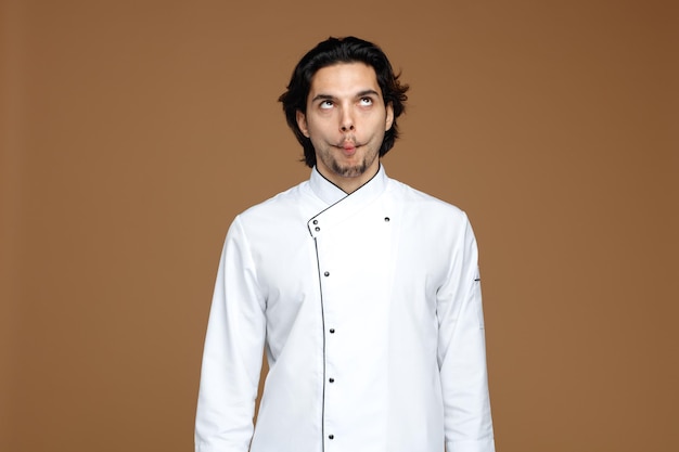 Free photo crazy young male chef wearing uniform looking up making fish face isolated on brown background