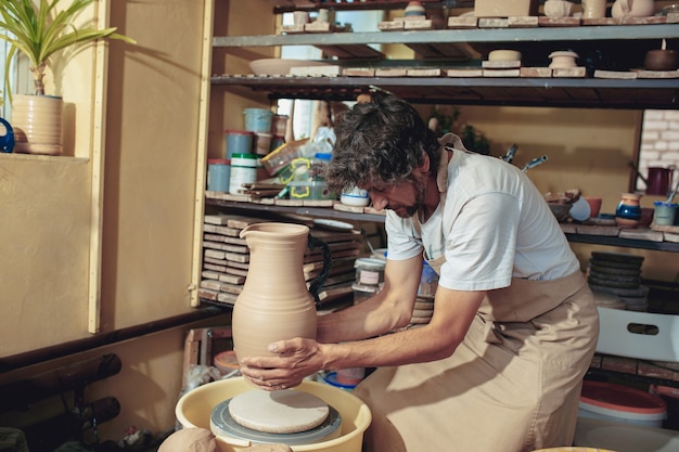 Creating a jar or vase of white clay close-up. Master crock. Man hands making clay jug macro. The sculptor in the workshop makes a jug out of earthenware closeup. Twisted potter's wheel.