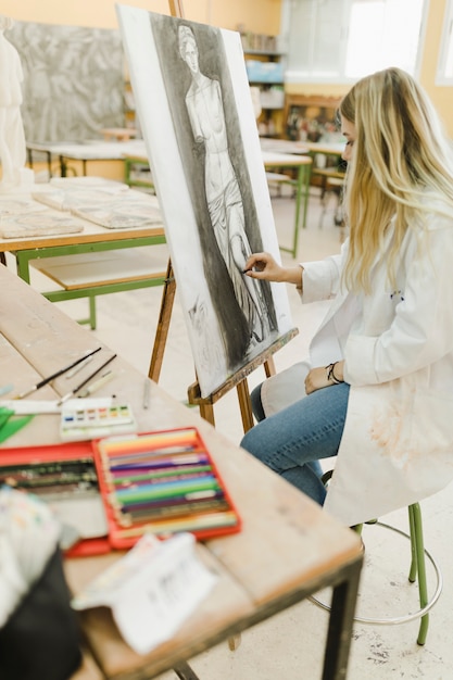 Free Photo creative artist sitting on stool sketching on easel with black crayon