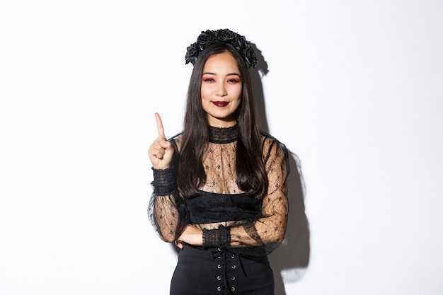 Free photo creative young woman in witch costume smiling pleased as have great idea, raising finger to say suggestion. female asian dressed-up like widow or mysterious magician, white background.