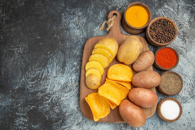 Crispy chips and uncooked potatoes on wooden cutting board and different spices on the left side of gray table