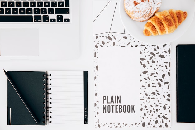 Croissant; plain notebook; laptop; pencil and spiral notepad on white desk