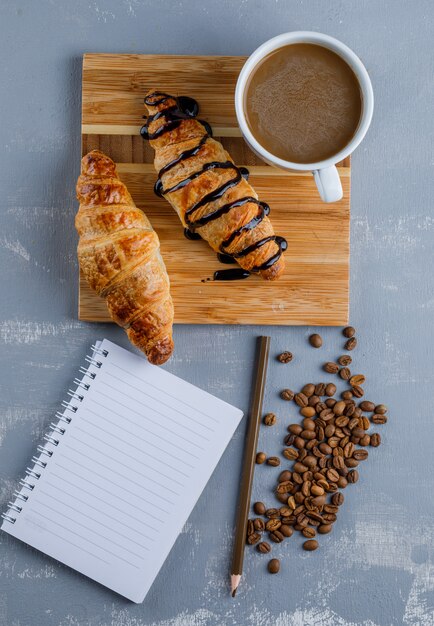 Croissants with coffee, notebook, pencil, coffee beans on plaster and wooden board, top view.