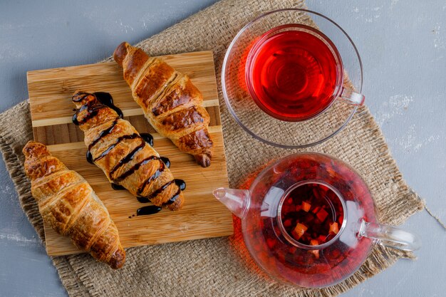 Croissants with sauce, tea, wooden board on plaster and piece of sack, flat lay.