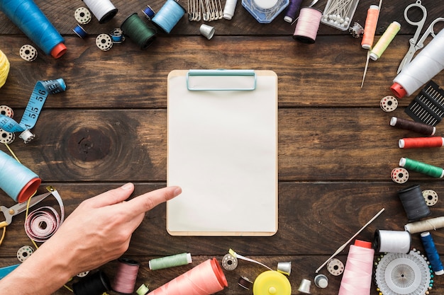 Crop hand pointing at empty clipboard near sewing stuff