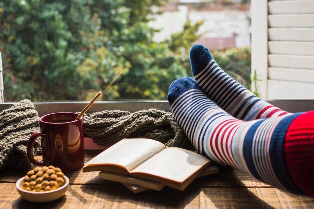 Crop legs near books and hot beverage