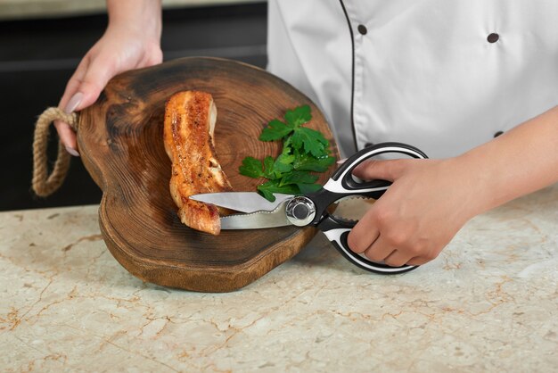 Cropped close up of a female chef cutting grilled meat with scissors working at the restaurant kitchen.