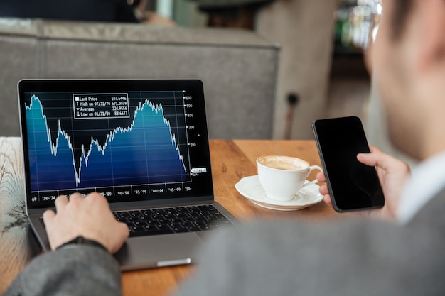 Free photo cropped image of businessman sitting by the table in cafe and analyzing indicators on laptop computer while using smartphone