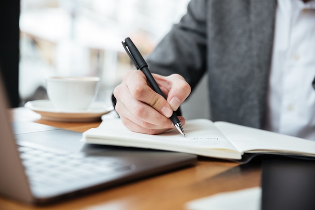 Cropped image of businessman sitting by the table in cafe with laptop computer and writing something