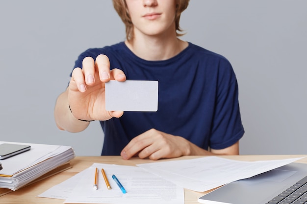 Free photo cropped image of casually dressed young male enterpreneur holds card with blnk copy space, sits on working table, sorrounded with papers, isolated over grey wall. businessman holds business card