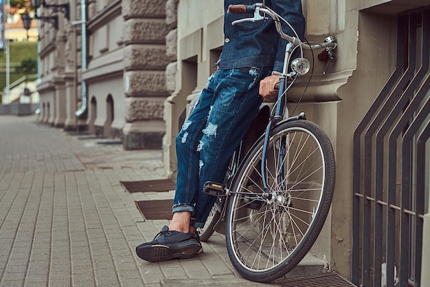 Cropped image of a fashionable man in stylish clothes leaning against a wall with city bicycle on a street.