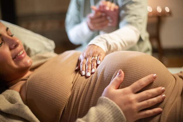 Cropped image of smiling pregnant woman and midwife at home. Woman in casual clothes lying on bed, midwife holding hand. Pregnancy, medicine, home birth concept