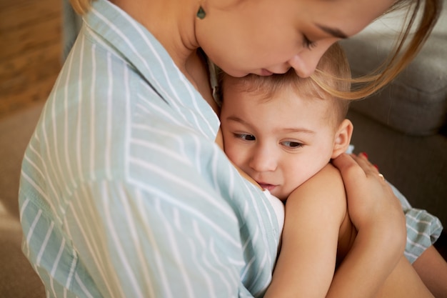 Cropped image of tender caring young mother craddling her sleepy baby in arms, singing song to him, kissing on forehead. Mom hugging one year old little child, trying to sooth abdominal cramps