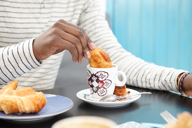 Free photo cropped image of unrecognizable afro american man dunking croissant into cup of cappuccino, enjoying delicious breakfast alone at coffee shop, sitting at table with mug and pastry. film effect