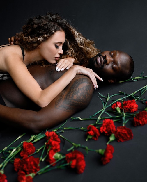 Free photo cropped of passionate woman with afro hairstyle wearing in bra lying on naked handsome tattooed afro american man with closing eyes near scattered red carnations