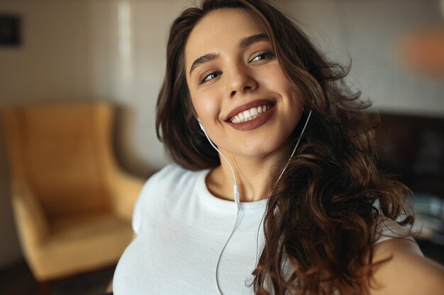 Cropped portrait of beautiful young European woman with long curly hair and brown lipstick wearing earphones looking with broad happy smile