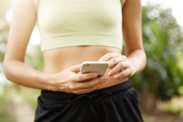 Cropped portrait of young female athlete using mobile phone during exercise outdoors.