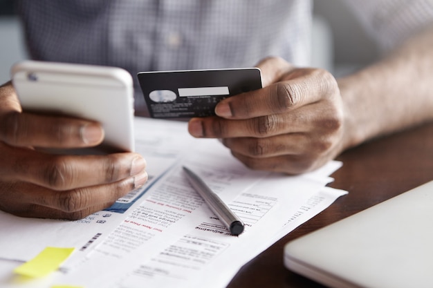 Cropped shot of African-American male holding cell phone in one hand and credit card in other