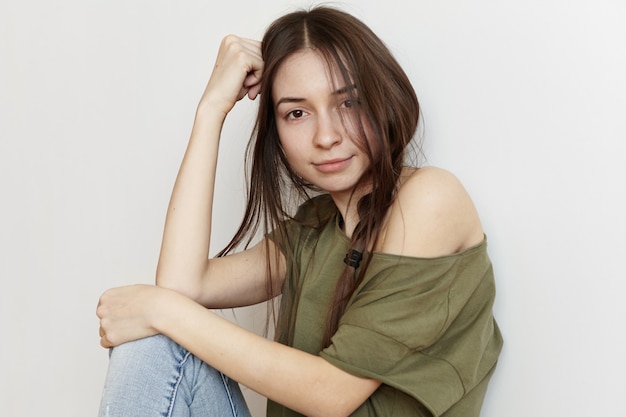 Cropped shot of fashionable beautiful young female with messy hairstyle wearing stylish clothes resting head on elbow and smiling, one shoulder showing up out of her army green oversize t-shirt