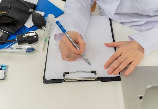 Cropped shot of female doctor hands working at desk with medical tools and laptop writing prescription on clipboard